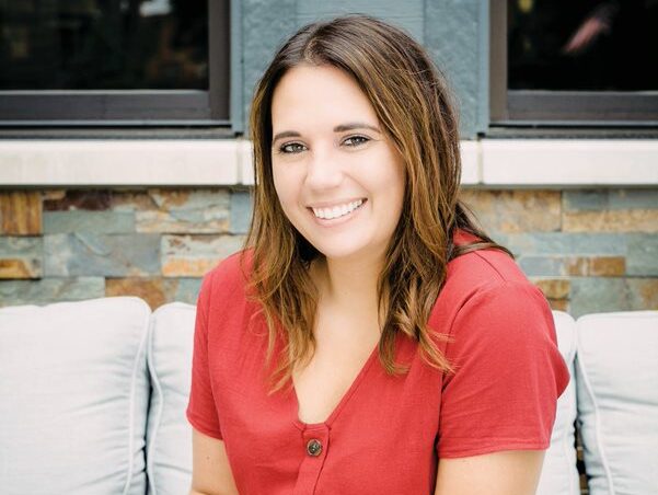 Sara Sybert sits outside on a patio couch in a short-sleeve red top