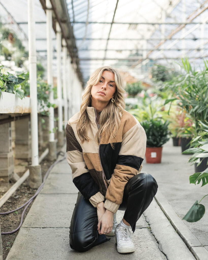 Trendy blonde woman in a patchwork natural toned jacket and jeans poses in a crouching position in a greenhouse nursery