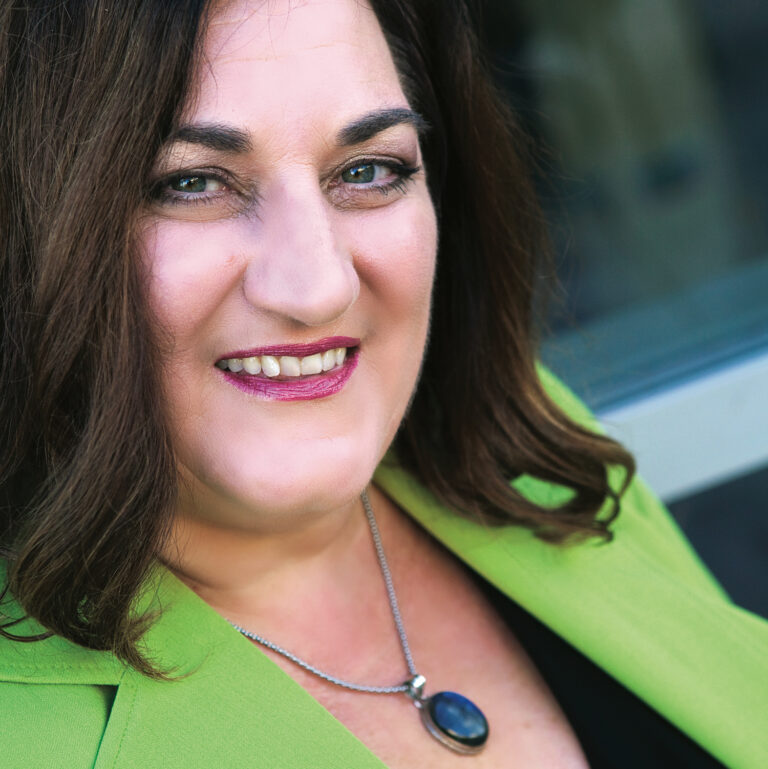 Liz Kitchell, president of SheMoolah, smiles at the camera wearing a lime green blazer and blue pendant necklace